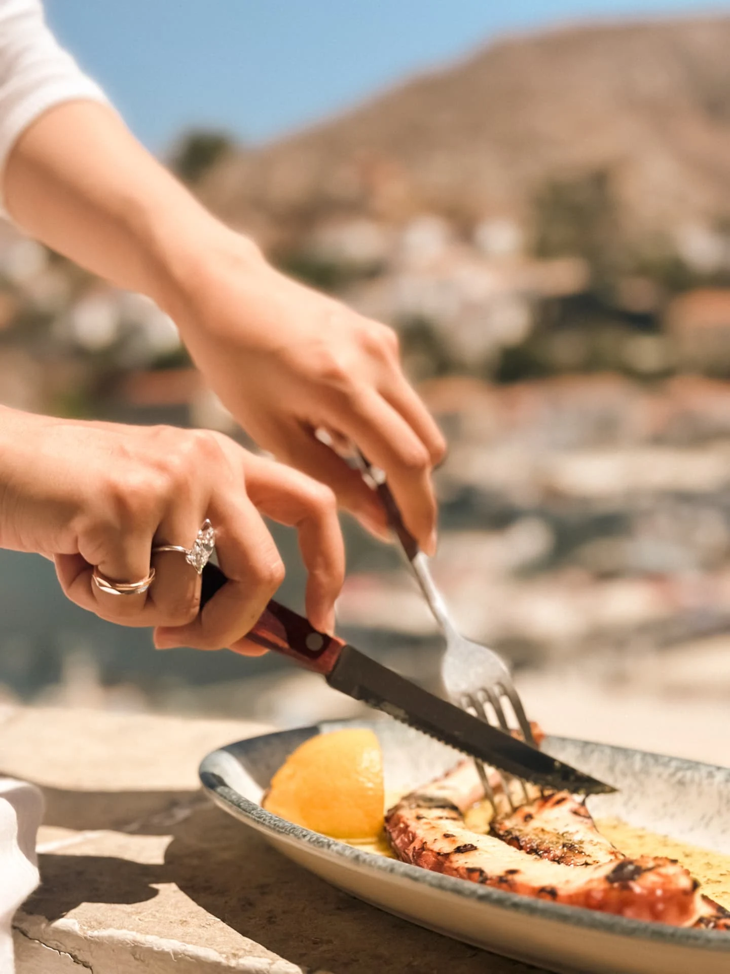 Hands cutting grilled octopus over a hillside town, rings catching the light